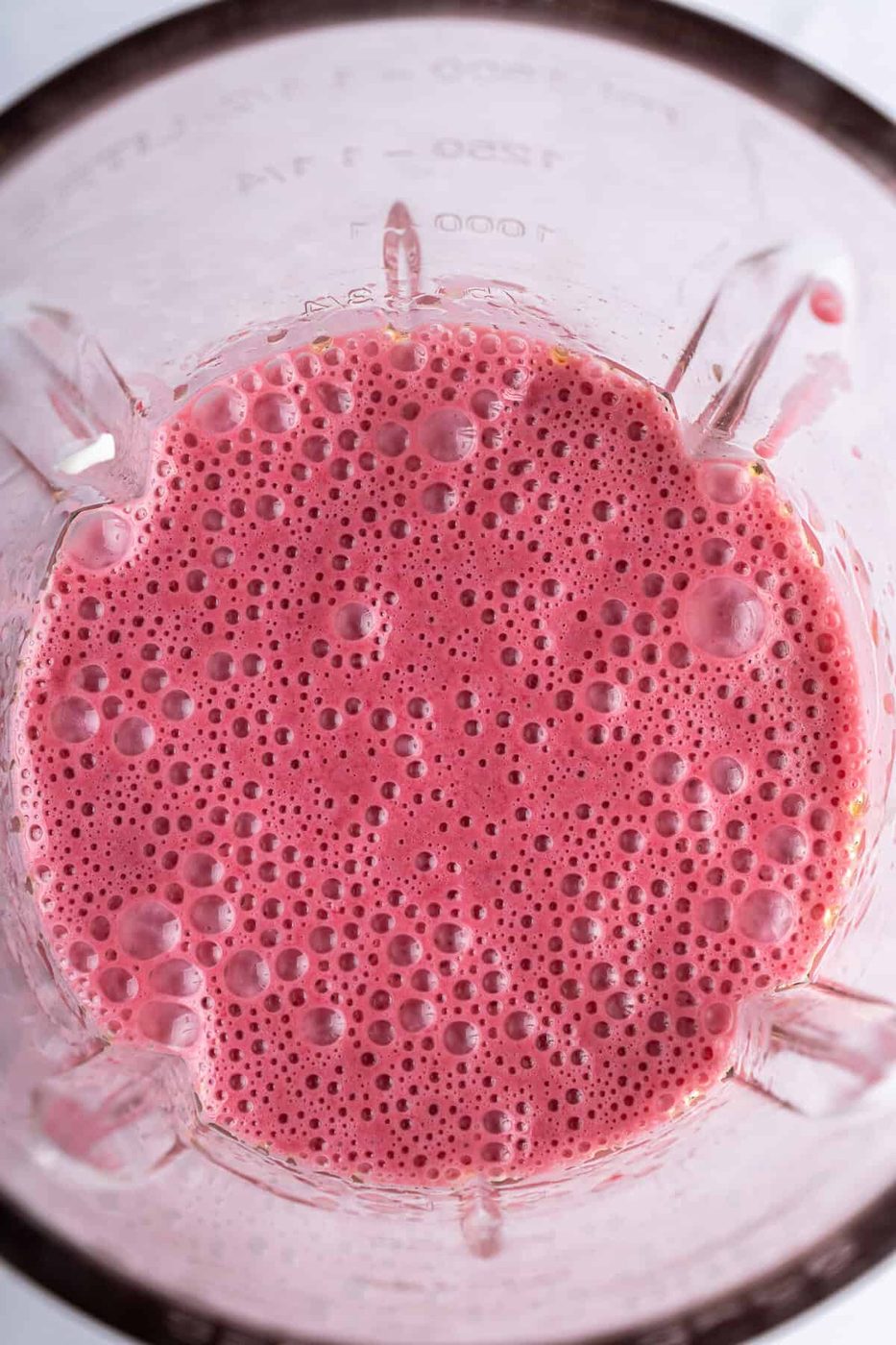 A blender filled with a frothy, pink smoothie. The surface shows a bubbly texture. The blender jar has measurement markings on the side.