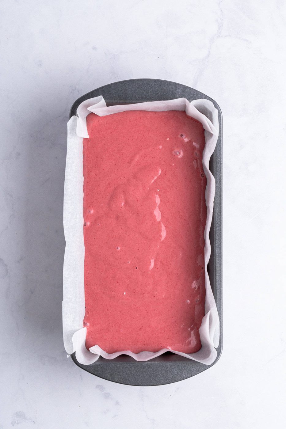 A loaf pan lined with parchment paper is filled with pink batter, ready for baking. The pan is set against a light marble surface.