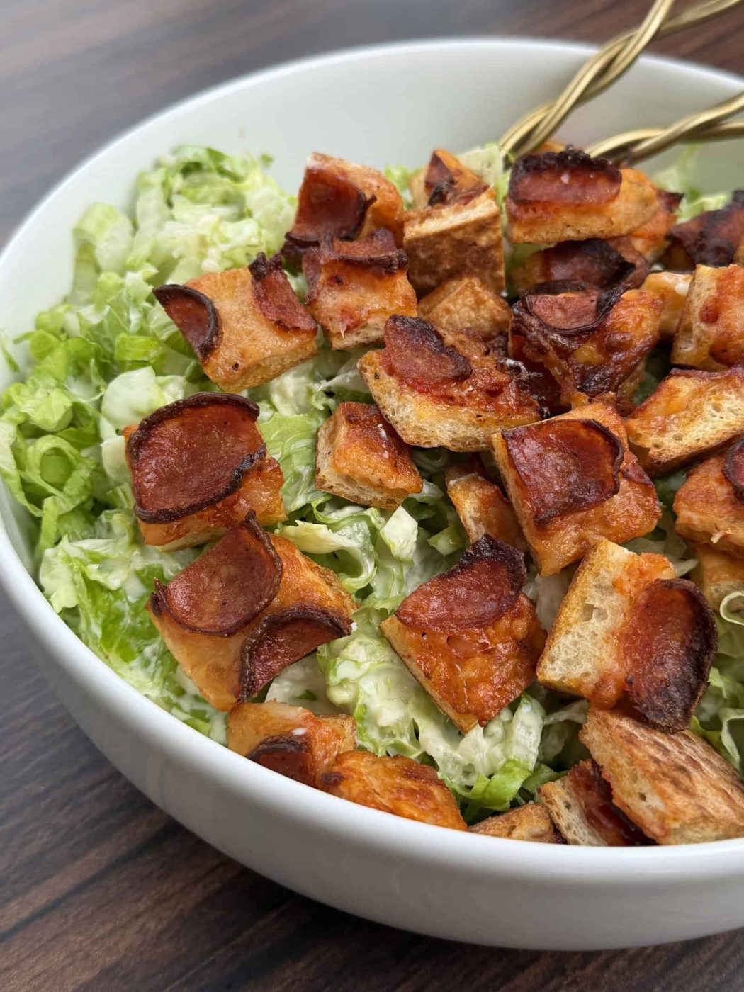 A close-up of a Pizza Crouton Salad in a white bowl, topped with crispy, browned croutons and diced vegetables on shredded lettuce. Two gold utensils rest inside the bowl on a wooden surface.