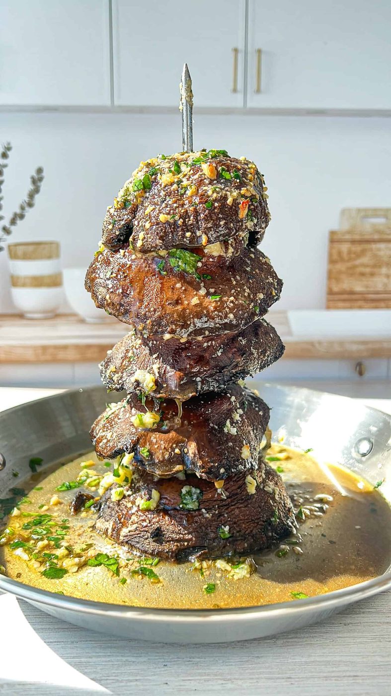 A stack of grilled meat chunks skewered vertically in a pan with sauce. The meat is garnished with herbs and grated cheese. Sunlight illuminates the dish, and the kitchen background is softly blurred.