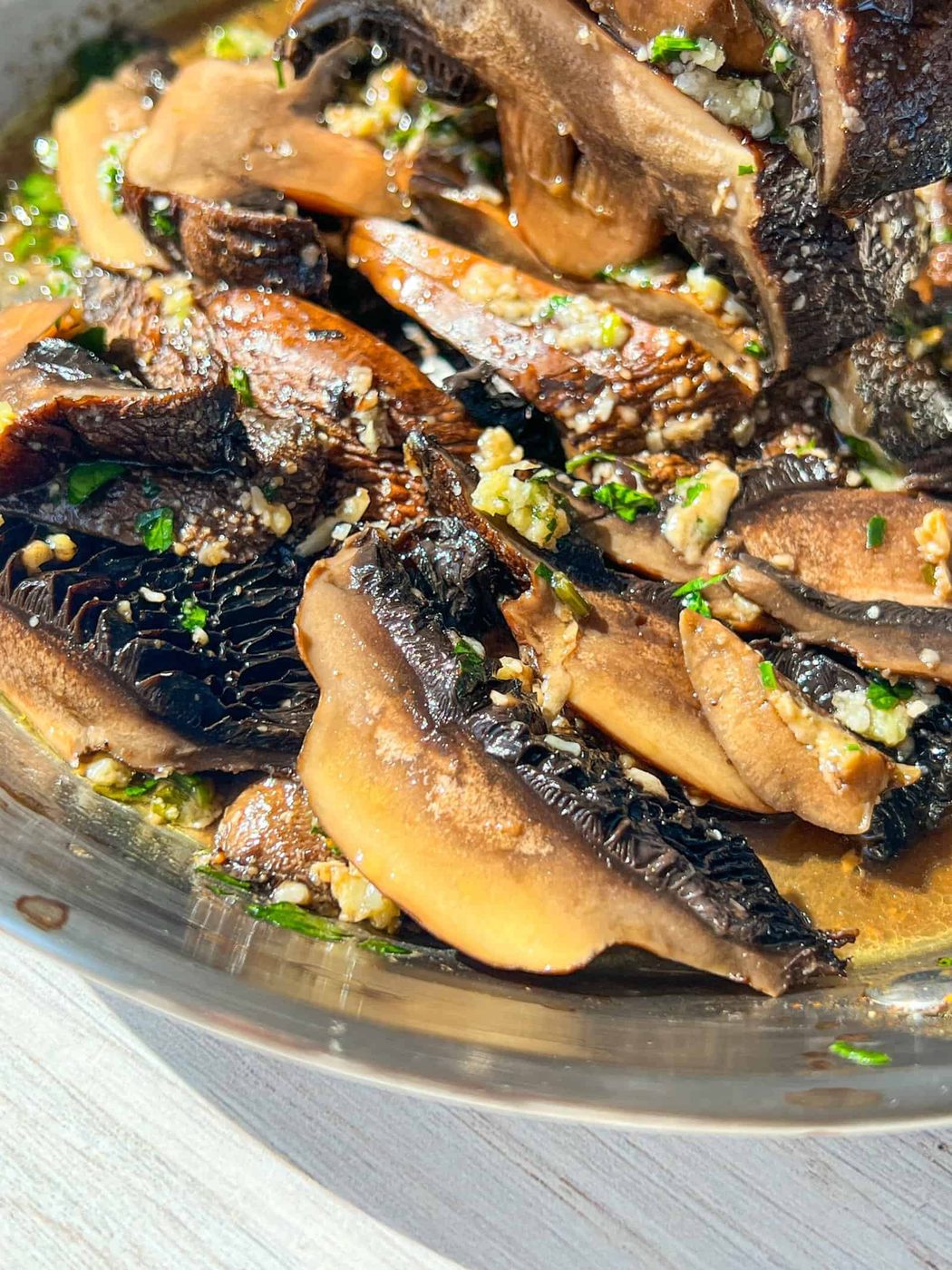A close-up of sautéed mushrooms in a dish, glistening with olive oil and garnished with minced garlic and chopped herbs. The mushrooms are a rich brown color, and the bright sunlight enhances their texture.