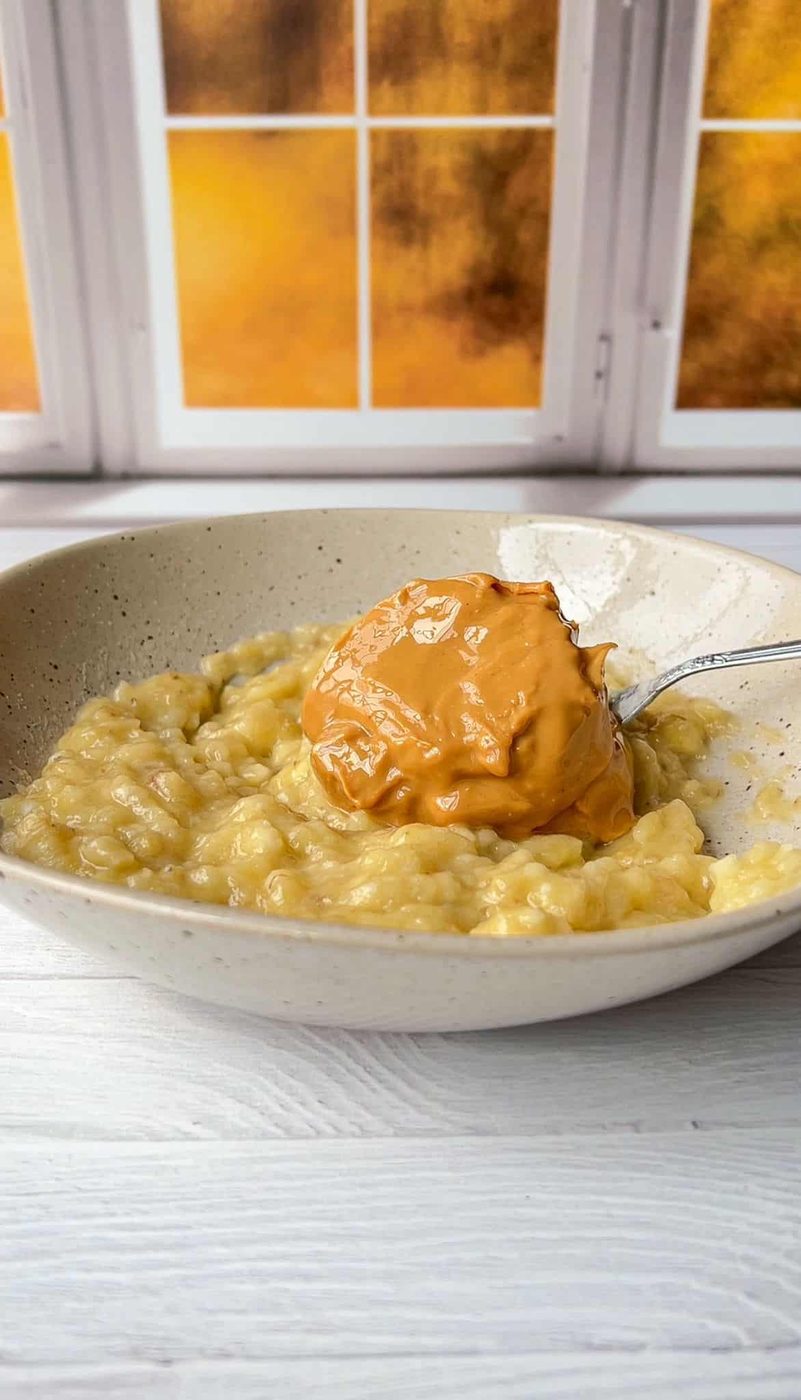 A bowl of mashed bananas with a spoonful of creamy peanut butter on top. The dish is set against a white wooden background with a window showcasing a warm, golden sunset view.