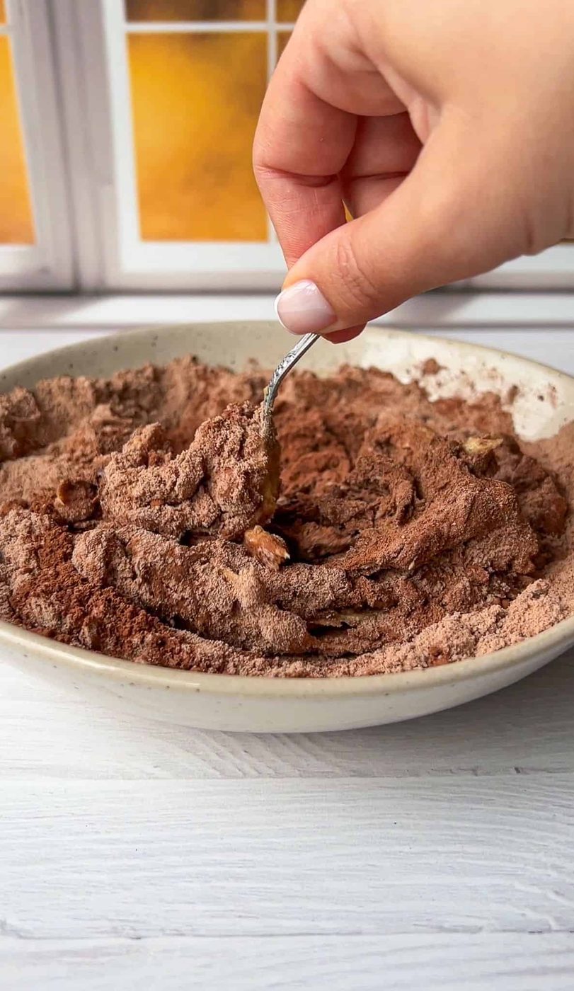 A hand holding a spoon mixes a bowl of cocoa powder and chopped nuts. The mixture is in a ceramic bowl on a light-colored wooden surface, with a blurred window in the background.