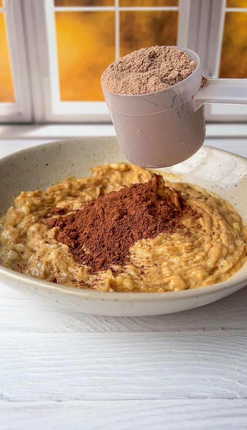 A bowl of oatmeal topped with a heap of cocoa powder. A hand holds a scoop of cocoa powder above the bowl, ready to add more. The background features a window with a warm, sunlit view.