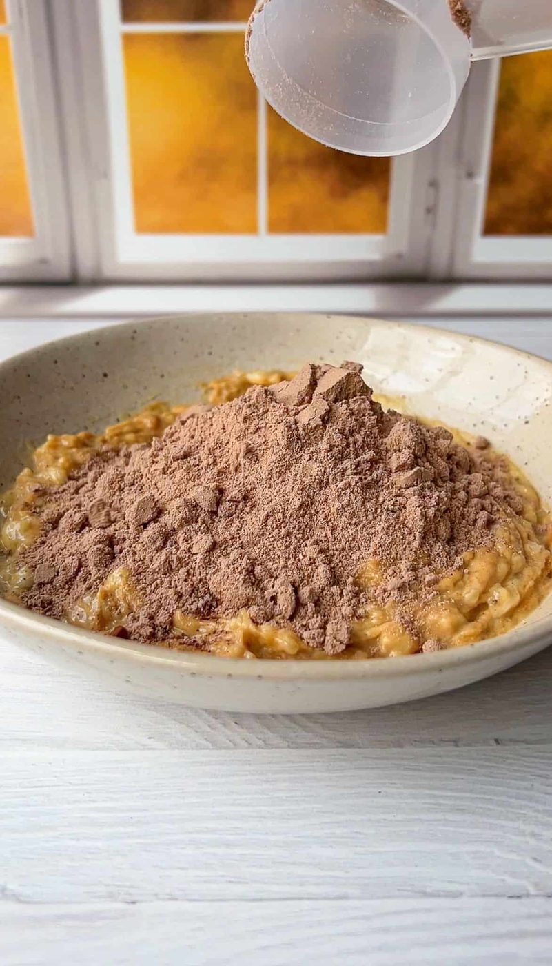 A bowl of oatmeal topped with a mound of cocoa powder on a light wooden table. A sifter is pouring more cocoa powder onto the oatmeal. A window in the background lets in warm, golden light.