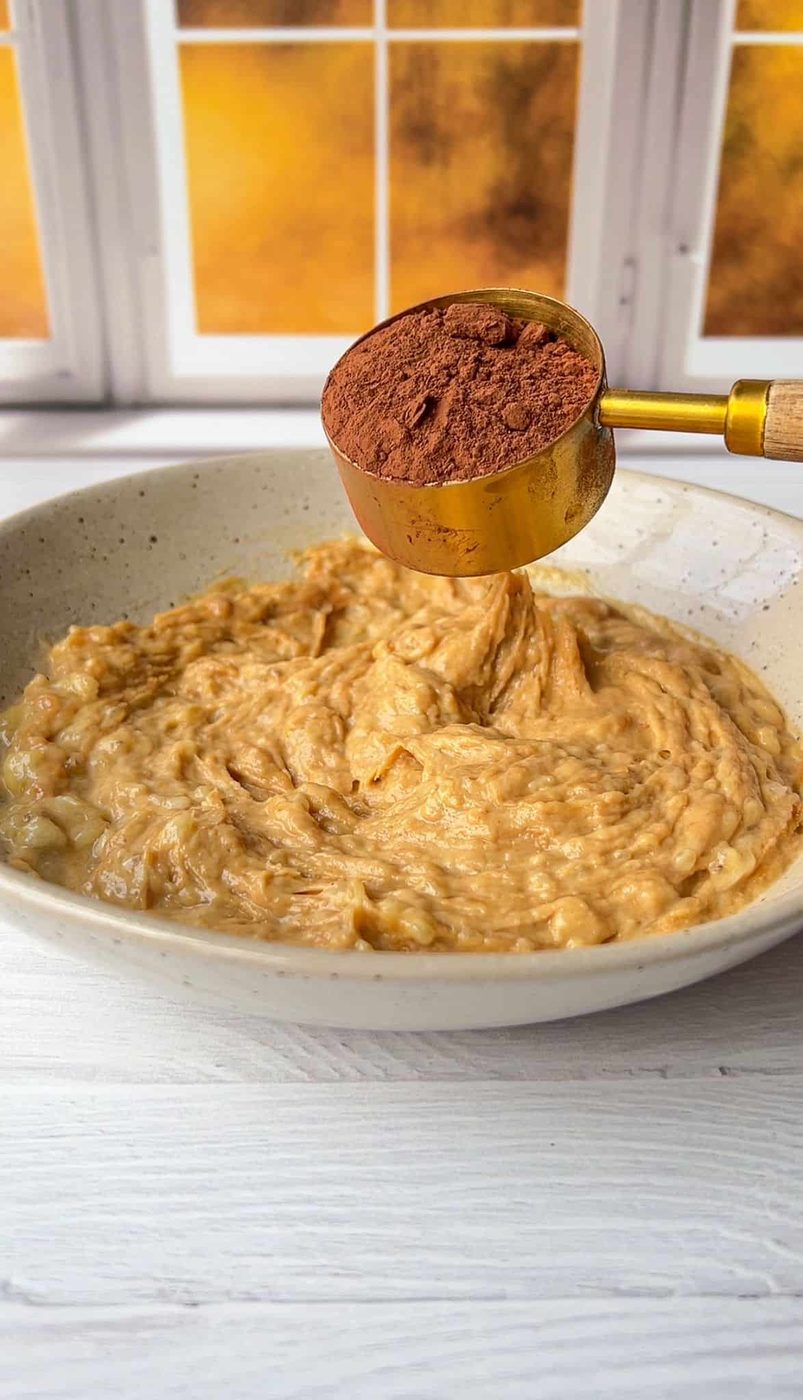 A bowl of mashed bananas sits on a white wooden table. Above the bowl, a golden measuring cup holds cocoa powder, ready to be added. The background features a window with a warm, sunny view.