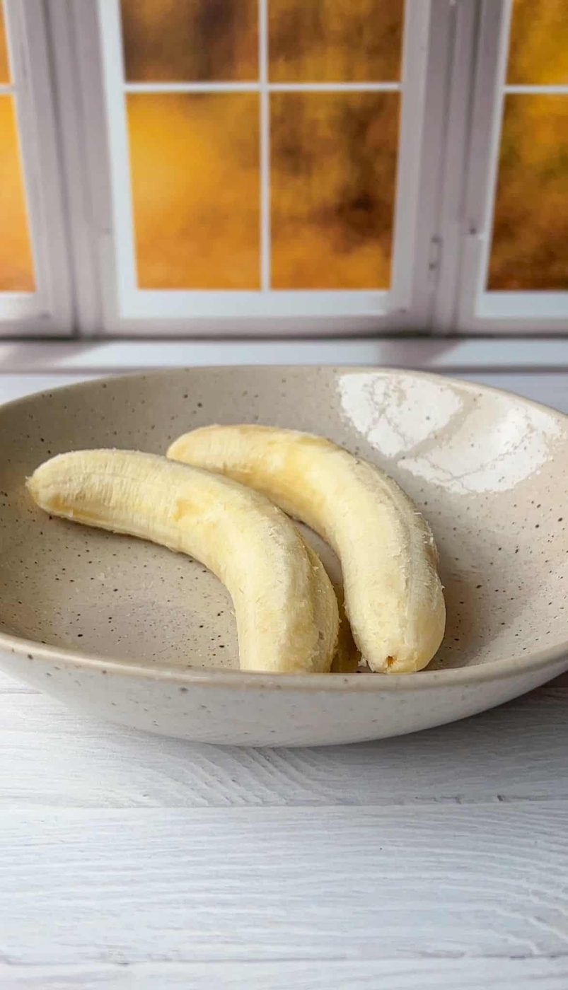 Two peeled bananas rest on a speckled ceramic plate. The plate is on a light-colored wooden surface, and in the background are windows showing a warm, blurred orange glow.