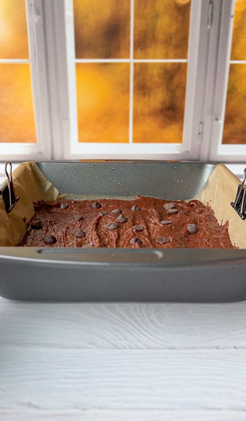 A baking pan with chocolate batter and chocolate chips on top is placed on a light-colored wooden surface. The pan is lined with parchment paper held by metal clips. A window with a soft orange glow is in the background.