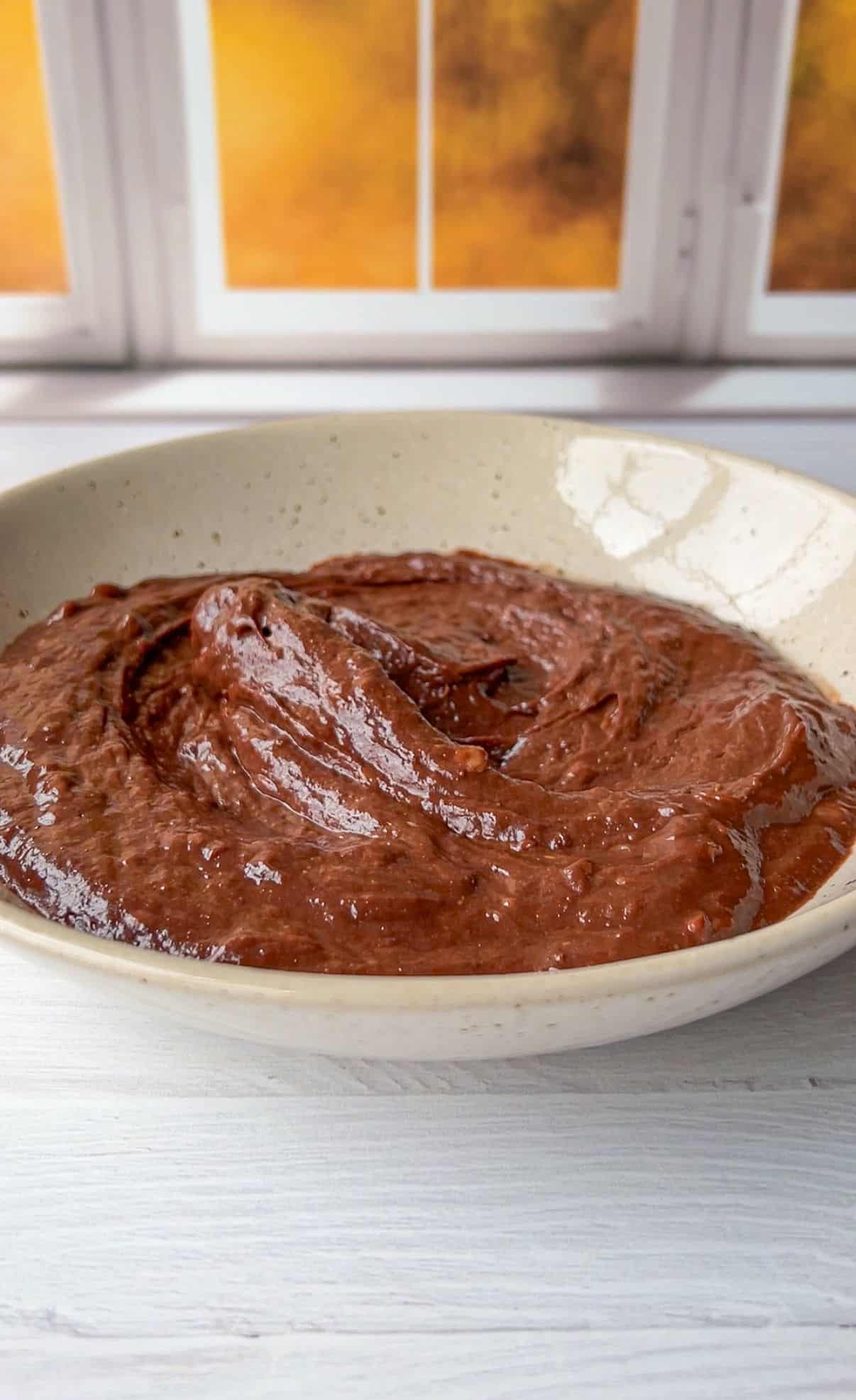 A bowl of thick, creamy chocolate pudding sits on a white wooden table. The smooth surface of the pudding glistens under natural light. A window with a warm glow is in the background.
