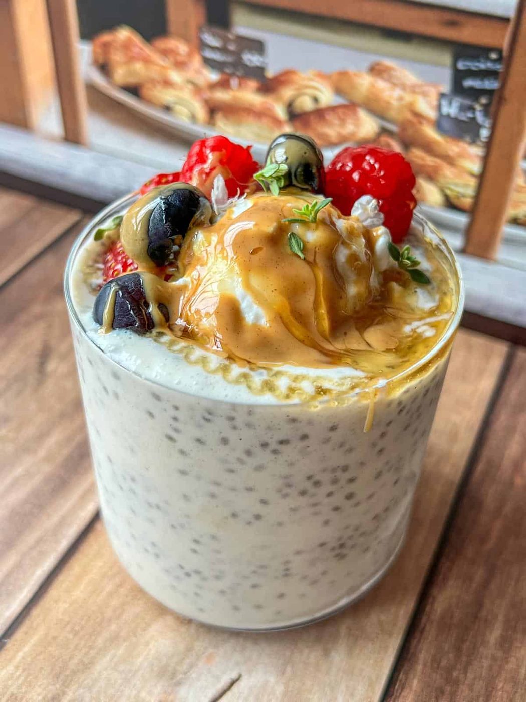A glass of chia pudding topped with berries, blueberries, peanut butter, honey, and fresh herbs, set on a wooden table with pastries visible in the background.