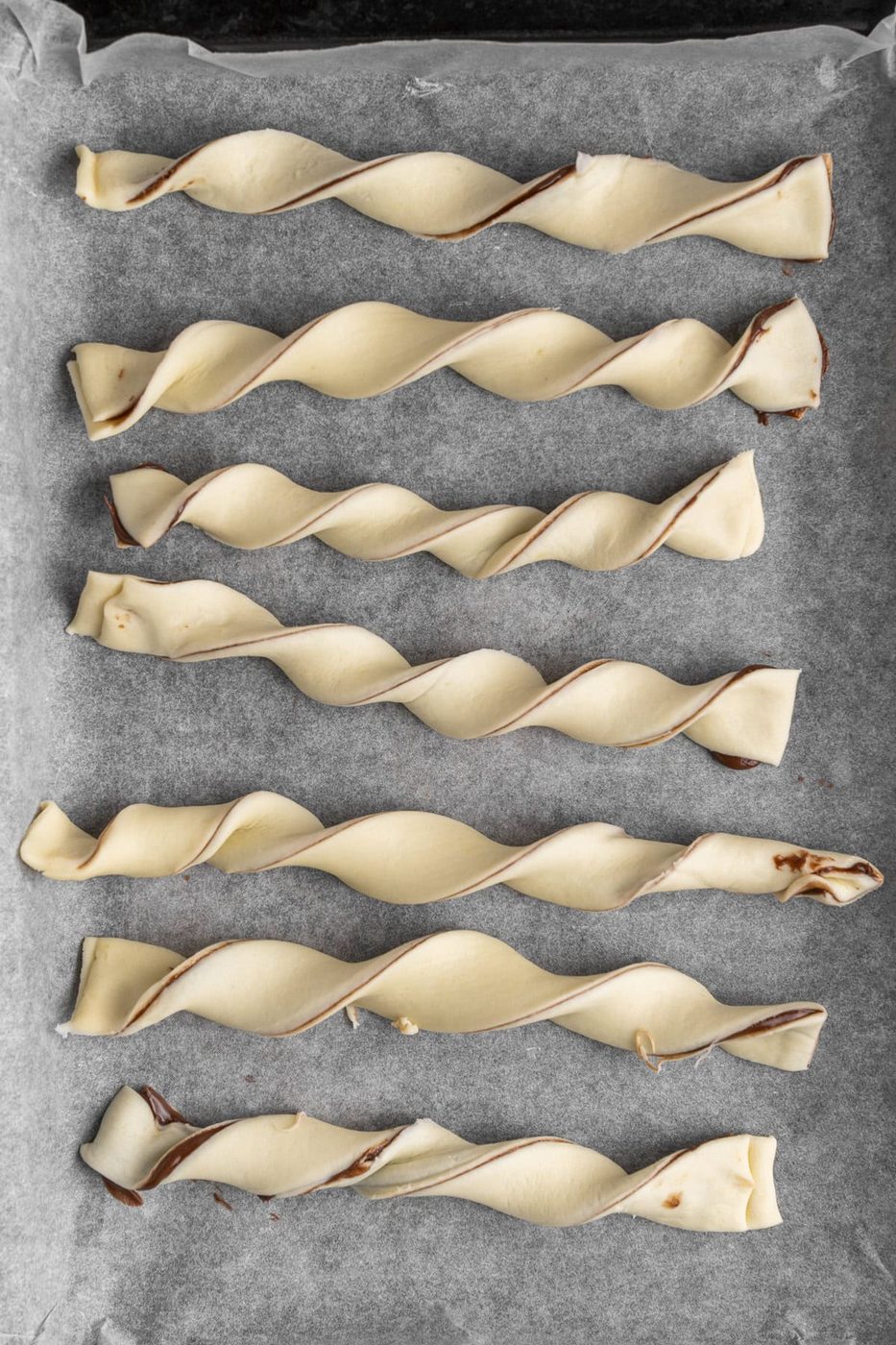 Seven uncooked twisted pastry sticks with chocolate filling are laid out on parchment paper on a baking tray.