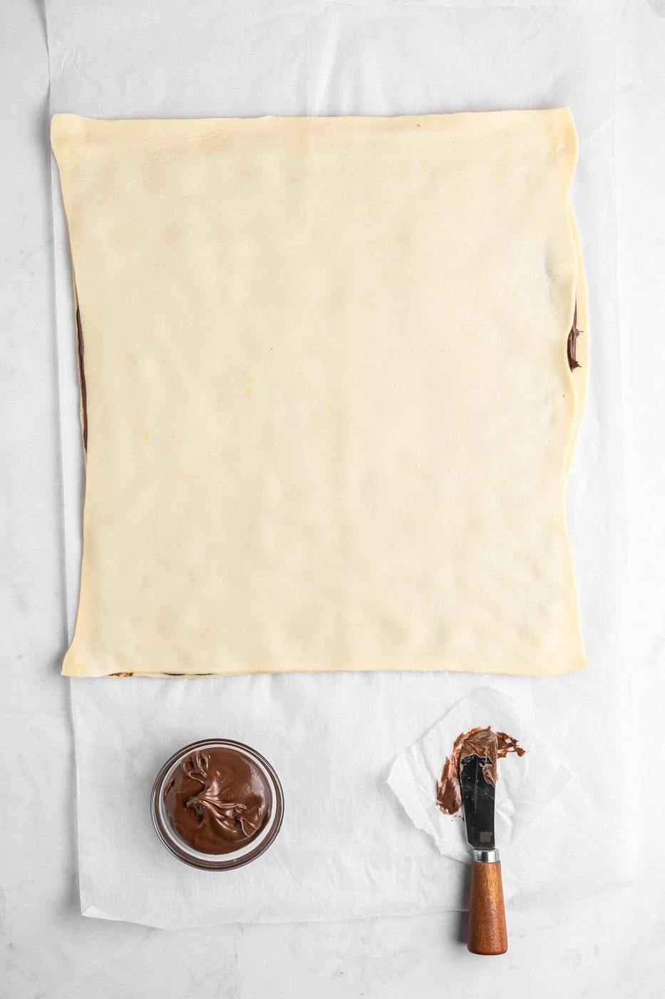 Square sheet of dough on parchment paper, partially spread with chocolate hazelnut spread. A jar of the spread and a wooden-handled spatula with some spread on it sit next to the dough.