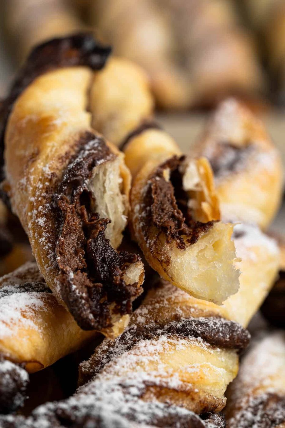 Close-up of flaky pastry twists filled with chocolate, dusted with powdered sugar. The pastries are arranged in a stack, showcasing their golden-brown exterior and rich chocolate layers.