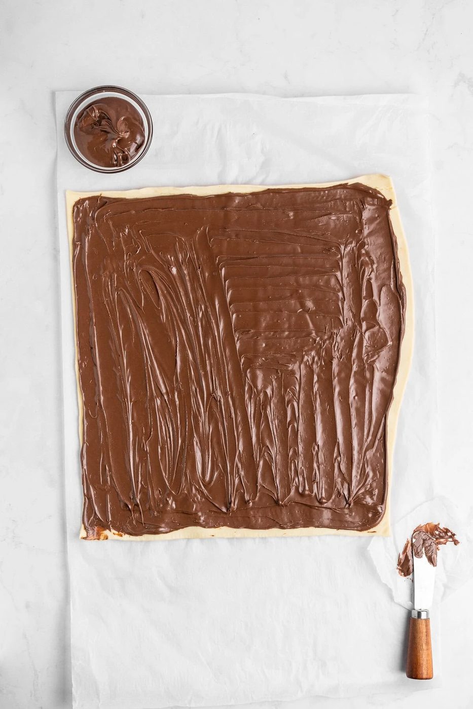 A sheet of dough lies on parchment paper, generously spread with chocolate hazelnut spread. A small bowl containing more of the spread and a metal spatula with some of the spread on its blade are positioned nearby.