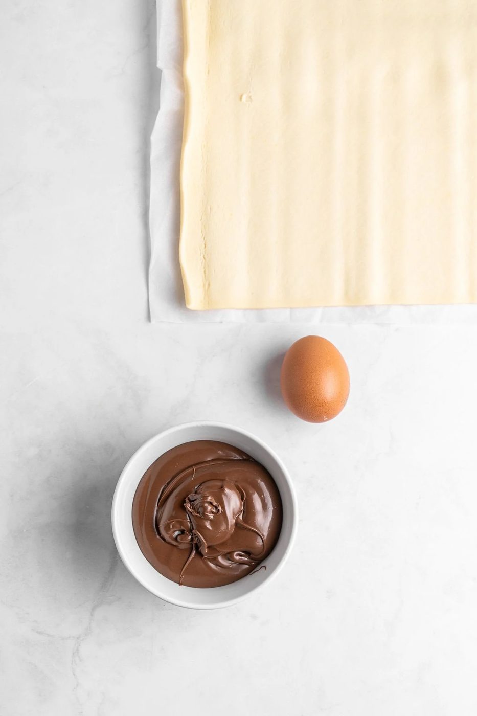 A sheet of puff pastry lies on parchment paper. Below it, there is a whole brown egg and a small white bowl filled with smooth chocolate spread, all placed on a light marble surface.