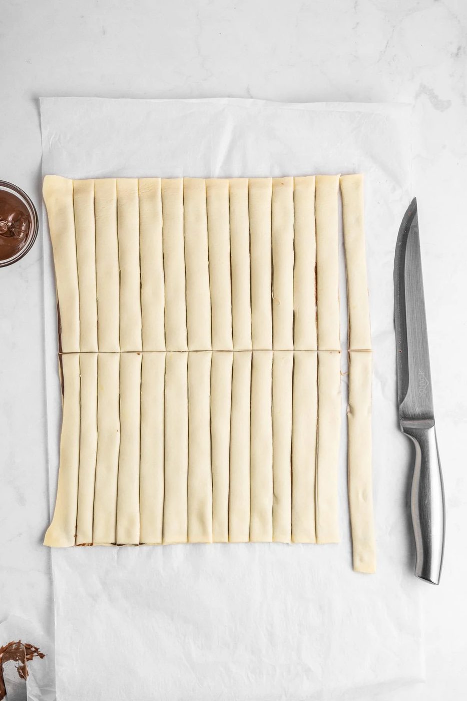 A sheet of dough is laid out on a surface, cut into evenly spaced vertical strips. A sharp knife rests beside the pastry, and a small container of chocolate spread is partially visible in the corner.