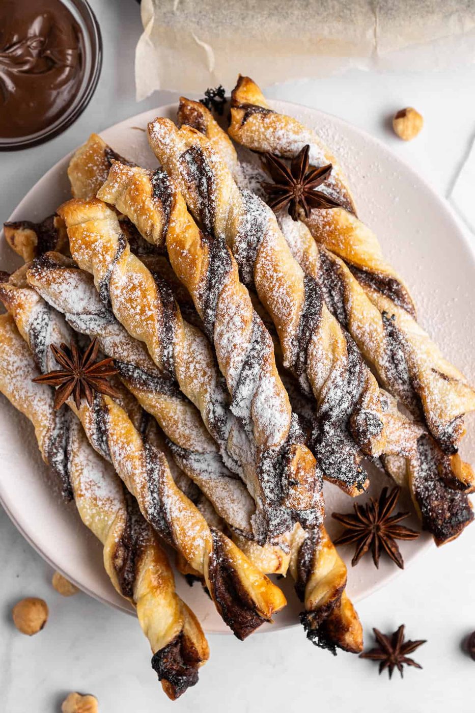 A plate of twisted puff pastry sticks with chocolate, dusted with powdered sugar. Star anise and hazelnuts are scattered around the plate. A small bowl of chocolate sauce is visible in the corner.
