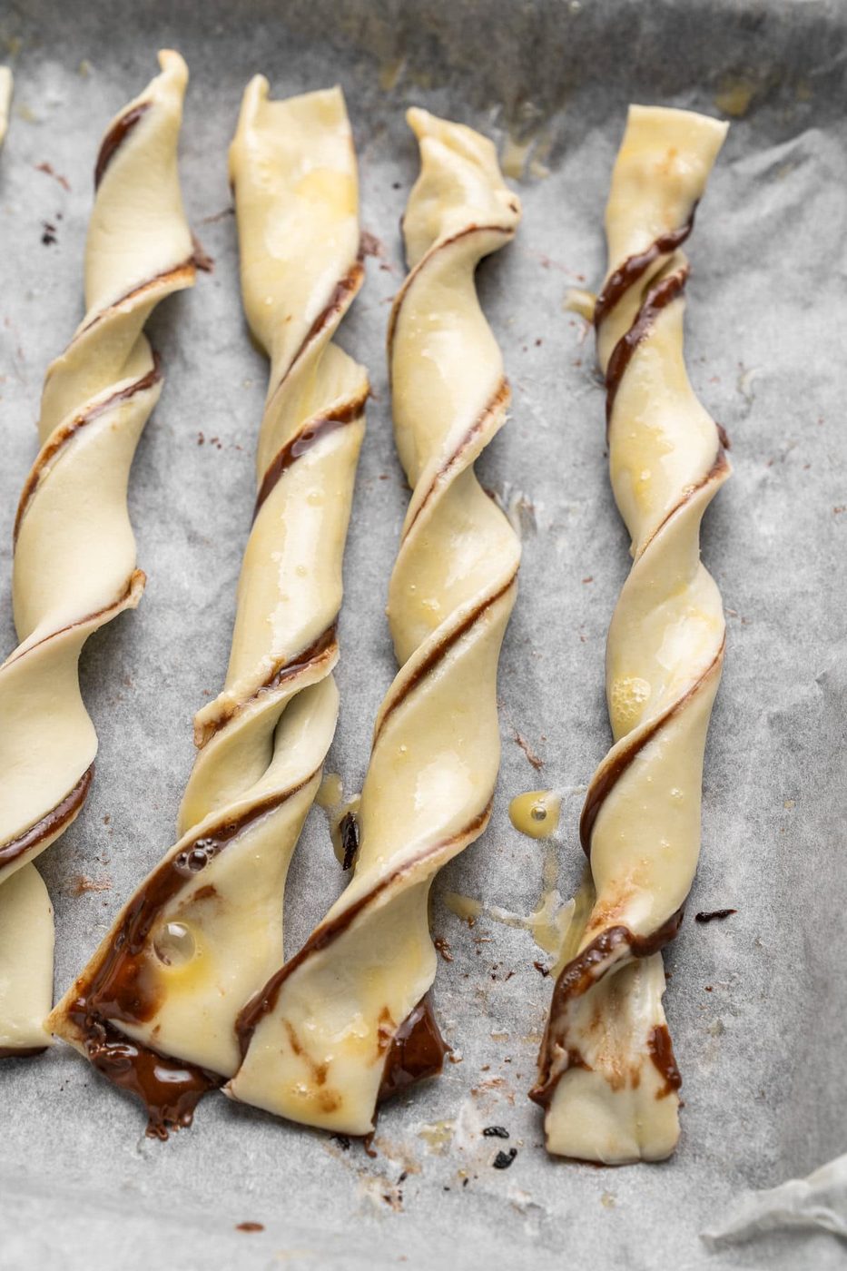 Four twisted pastry sticks with chocolate filling are arranged on a baking sheet lined with parchment paper. The pastries are slightly golden, with visible chocolate oozing from the twists.