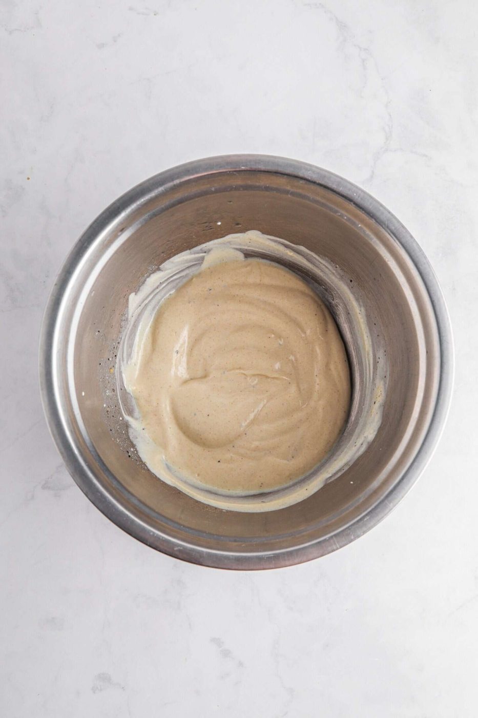 A stainless steel mixing bowl containing a smooth, light beige batter or mixture is placed on a white marble surface.