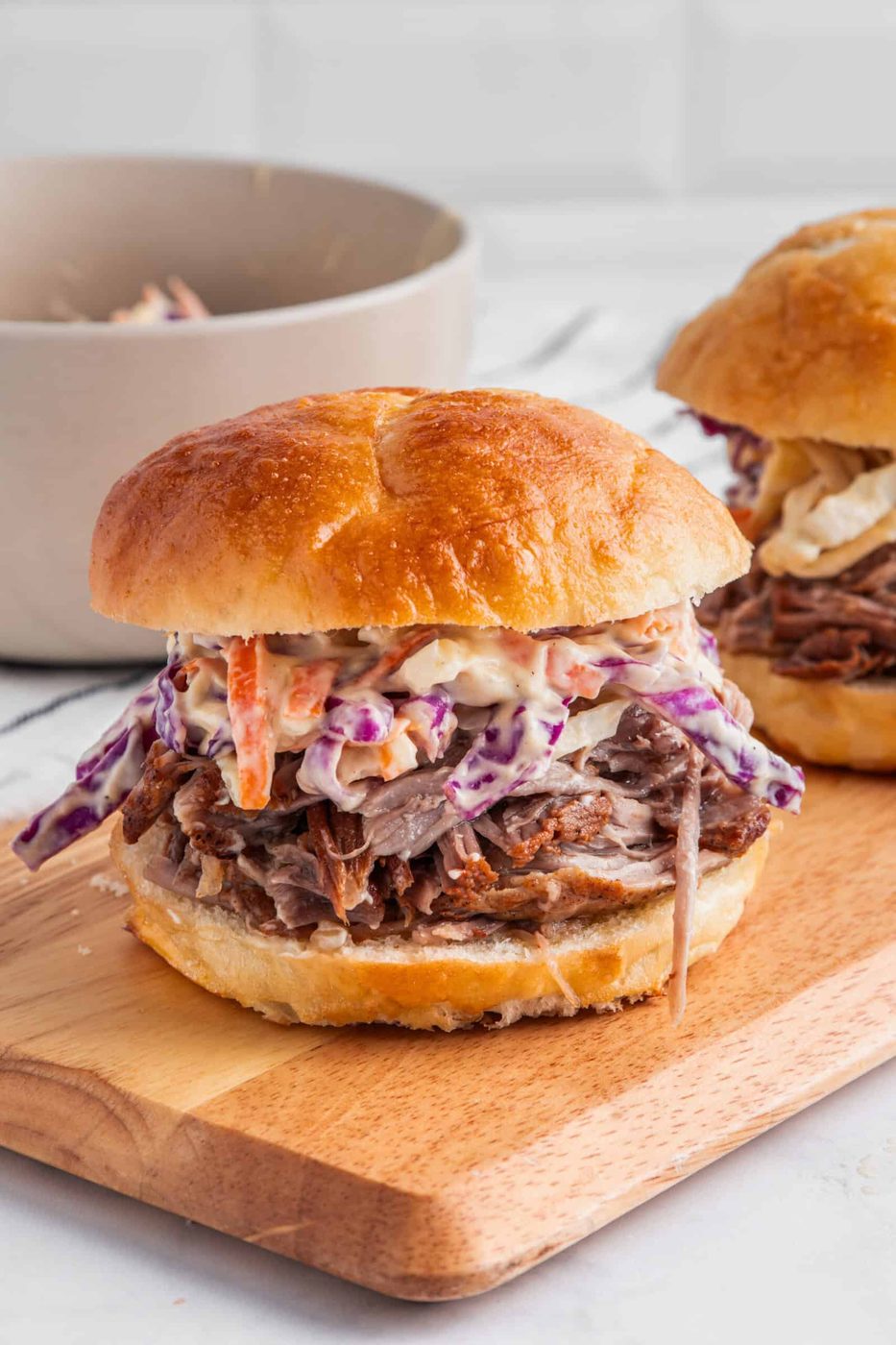 A pulled pork sandwich on a brioche bun is topped with creamy coleslaw, featuring red and white cabbage and carrots. Another sandwich is partially visible in the background, with a bowl on the left side, all on a wooden board.