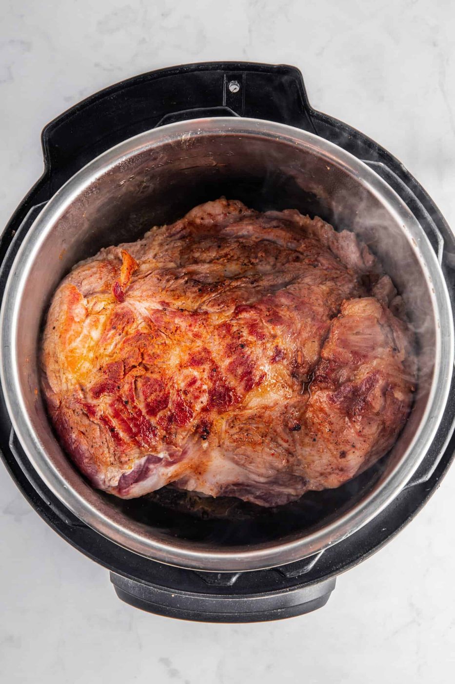 A large piece of browned meat sits inside an electric cooker on a marble countertop, with steam rising from it. The meat appears well-cooked with a crispy outer layer.
