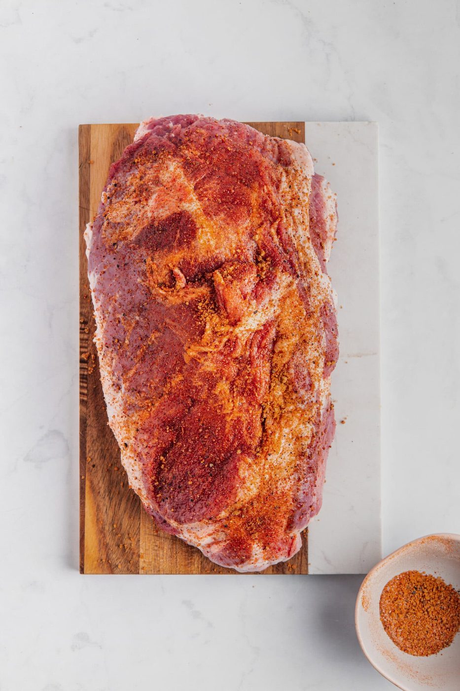 A raw piece of meat, seasoned with red spices, sits on a wooden and marble cutting board. A small bowl with more seasoning is nearby on the white, marble surface.