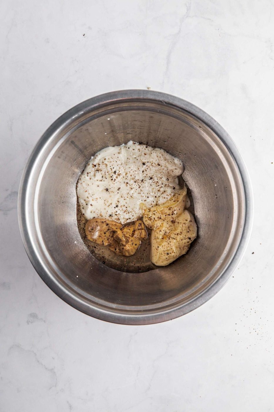 A stainless steel mixing bowl containing sour cream, mayonnaise, and a light sprinkle of ground black pepper, on a white marble surface.