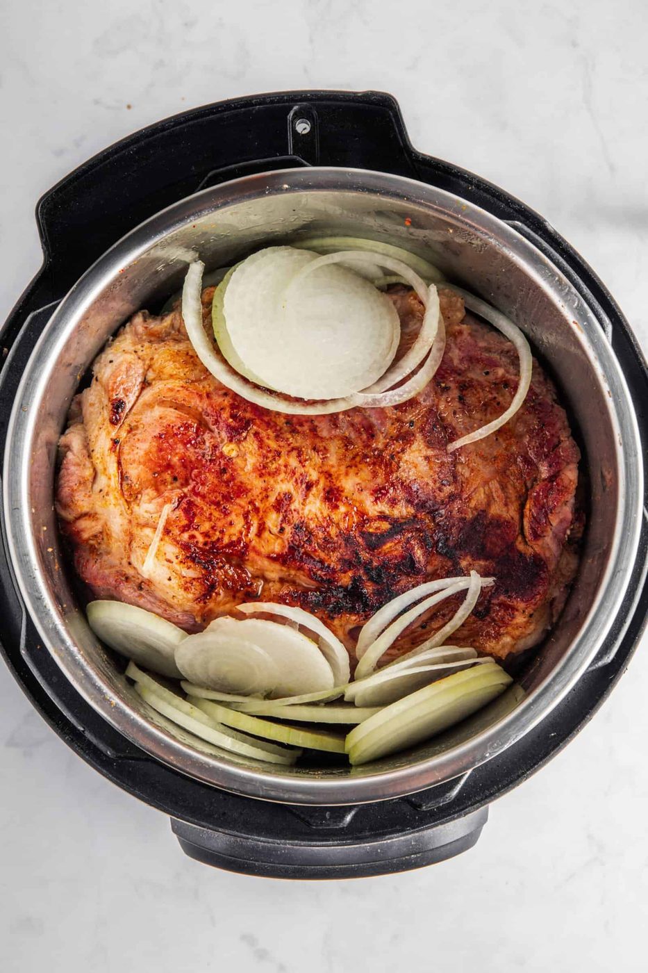 A seasoned piece of meat with sliced onions on top rests inside an instant pot on a marble countertop.