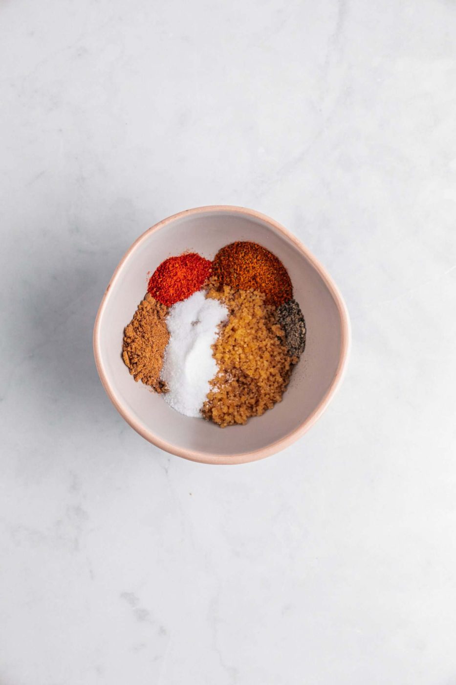 A bowl filled with various ground spices arranged in sections, including white, brown, red, and black colors, placed on a light gray marble surface.