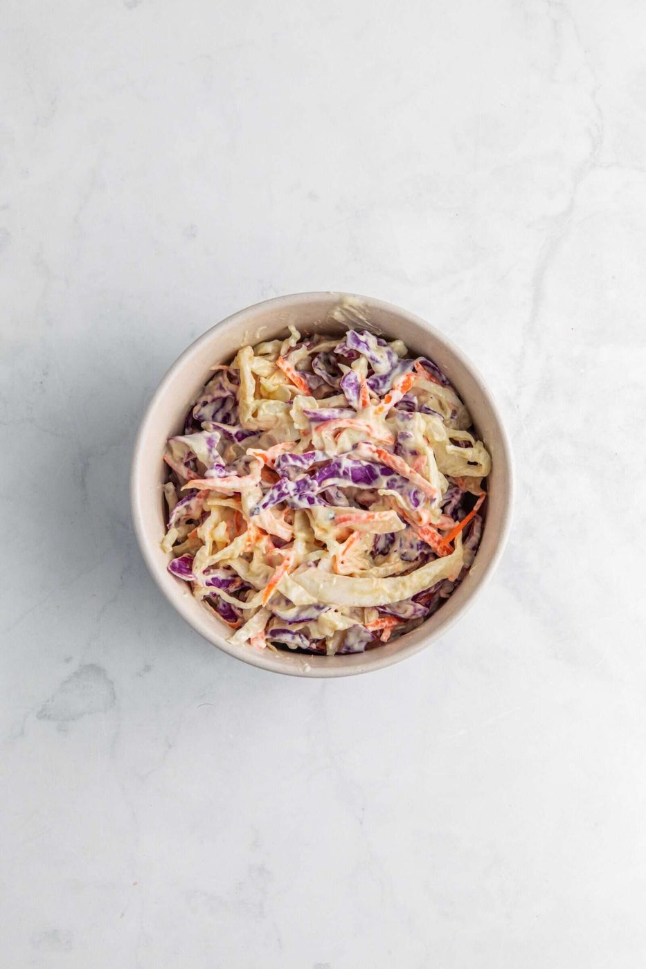 A bowl of creamy coleslaw, featuring shredded green and purple cabbage with orange carrot strips, set against a light marble surface.