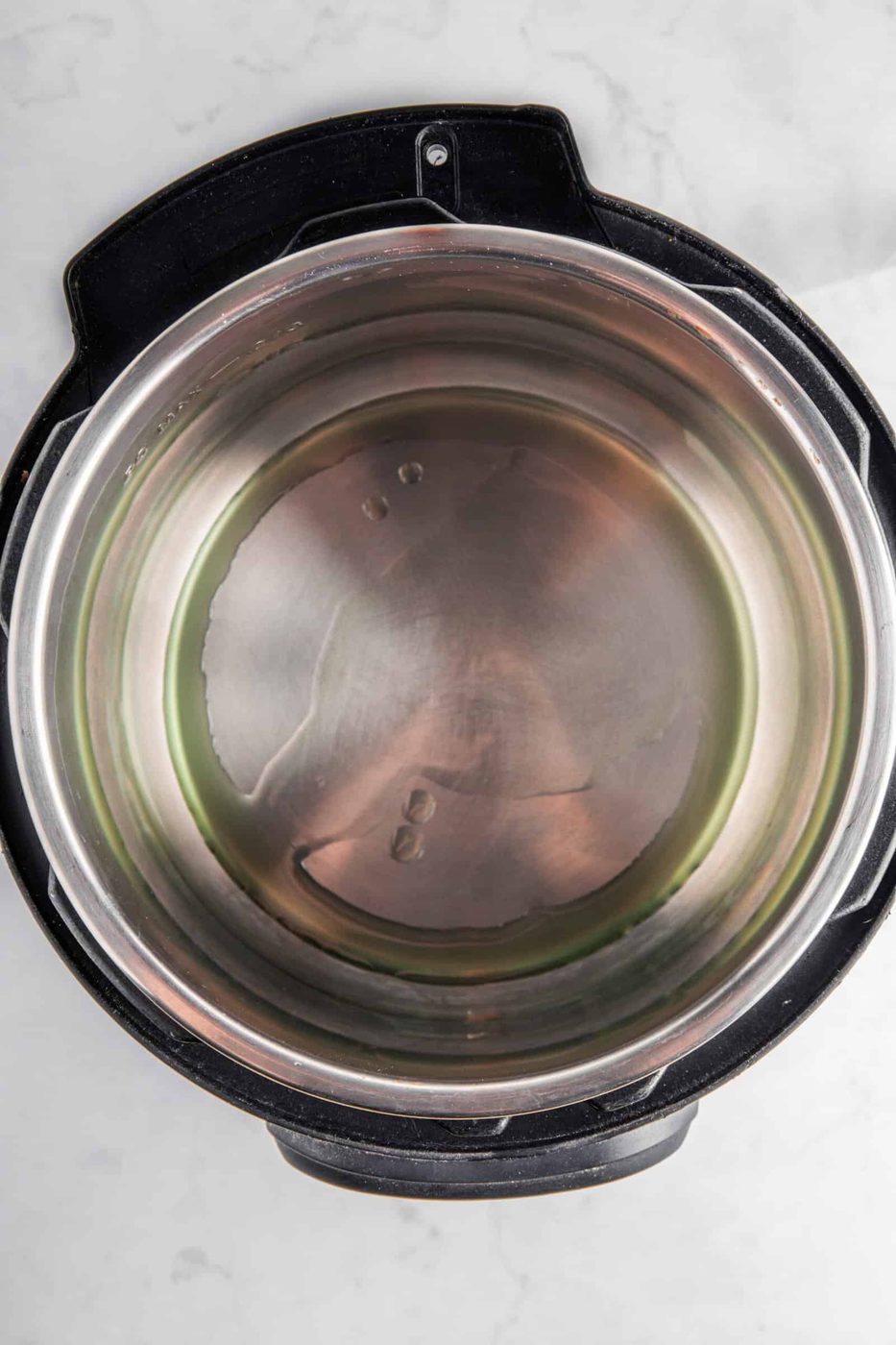 A stainless steel inner pot placed inside a black electric pressure cooker, viewed from above, on a white marble surface. The pot is empty and clean, with a reflective, slightly brushed finish.