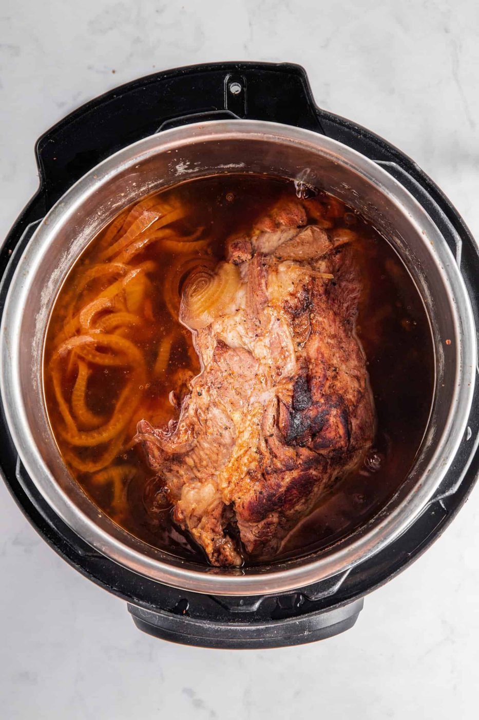 A seasoned piece of pulled pork meat with sliced onions on top rests inside an instant pot on a marble countertop.