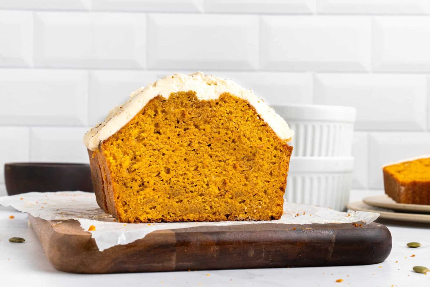 A loaf of pumpkin bread with white frosting sits on parchment paper atop a wooden cutting board, with a slice cut off and dishes in the background.