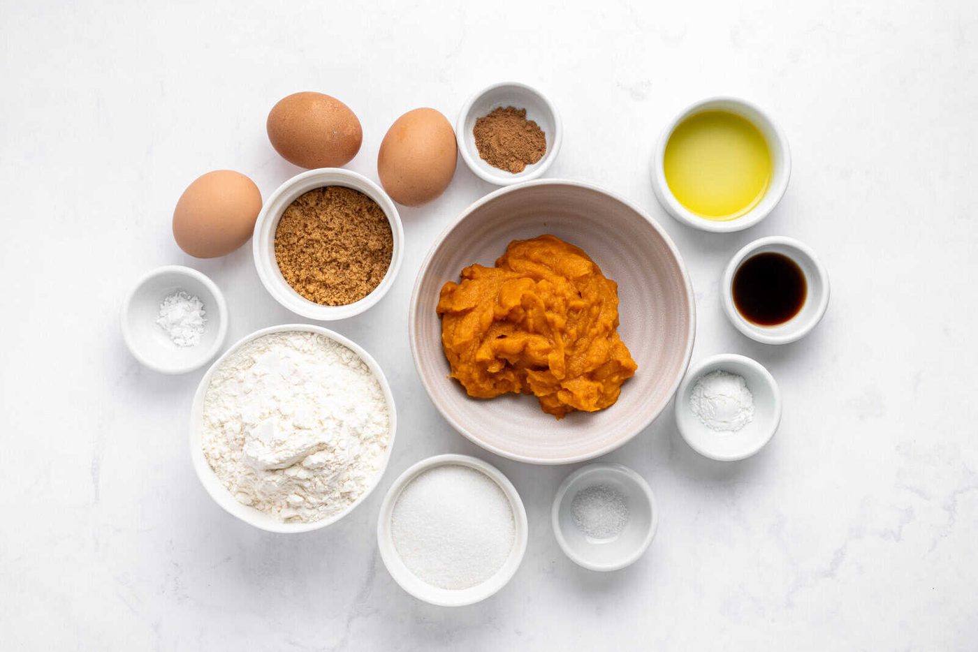 Bowls containing flour, sugar, brown sugar, baking powder, spices, salt, and vanilla surround a bowl of pumpkin puree. Three eggs and a small bowl of oil are also included, all arranged on a white surface.