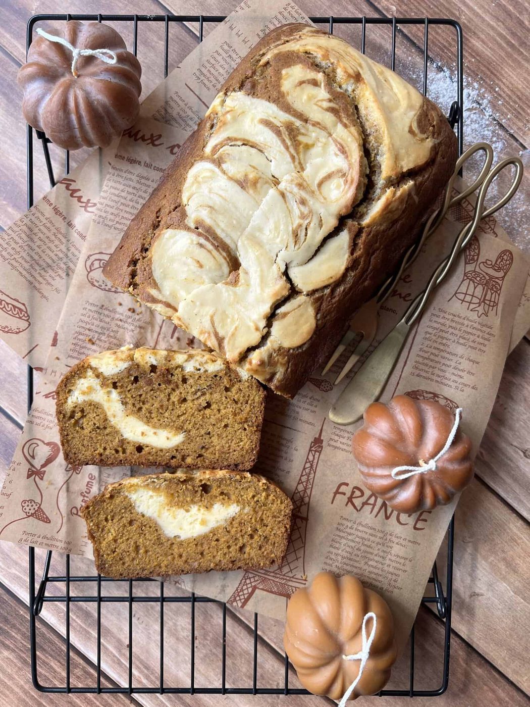 A loaf of pumpkin bread with cream cheese swirls, two slices cut, on a cooling rack with decorative paper, surrounded by three small faux pumpkins and two gold forks.