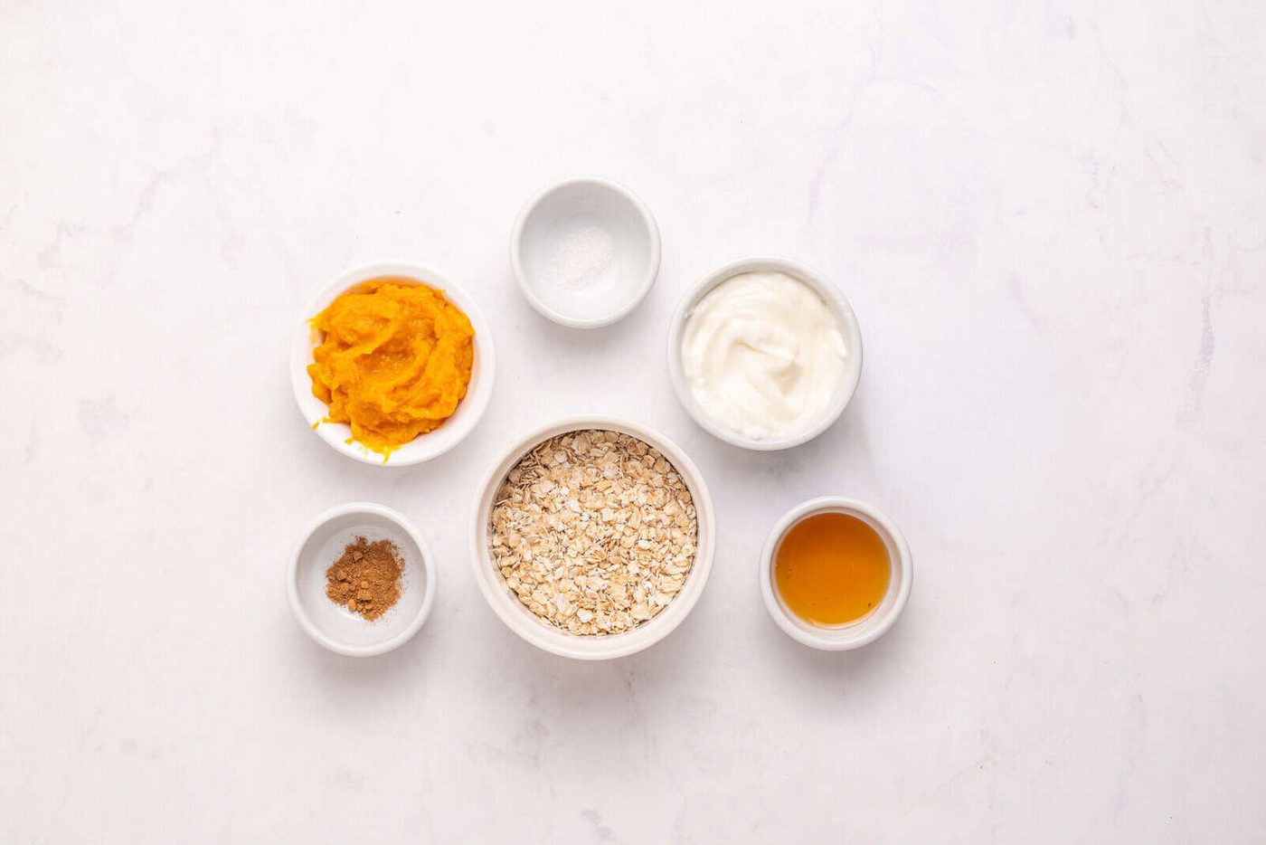 Six small bowls on a white surface, each containing a different ingredient: oats, pumpkin puree, yogurt, maple syrup, ground spice (possibly cinnamon), and a pinch of salt.