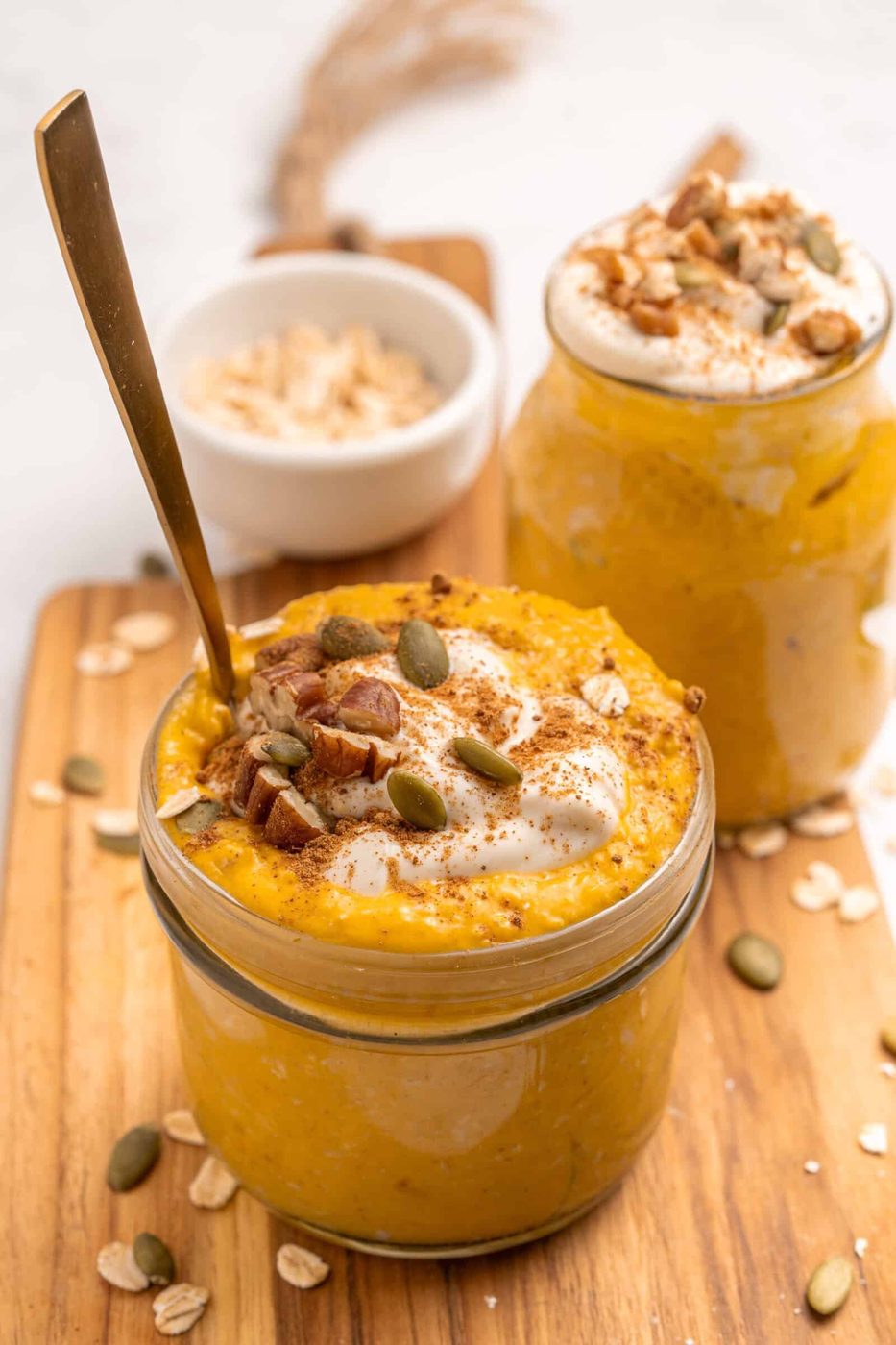 Two glass jars filled with creamy pumpkin overnight oats, topped with yogurt, pecans, pumpkin seeds, and a sprinkle of cinnamon. A spoon is in one jar. Rolled oats are scattered on the wooden board underneath.