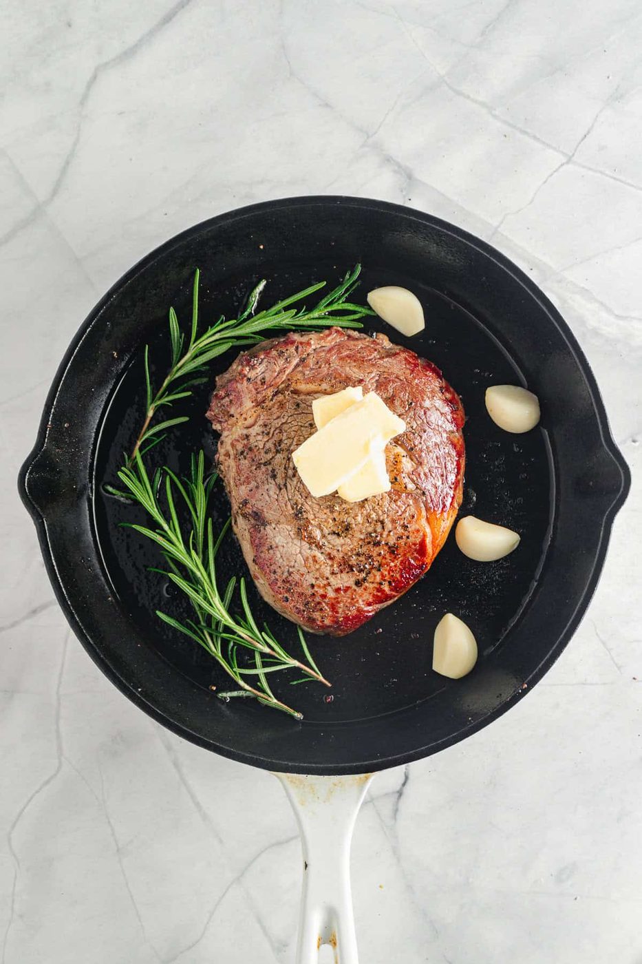 The seasoned meat topped with butter is in a black frying pan. Sprigs of rosemary and garlic cloves surround it. The pan sits on a light marble surface.