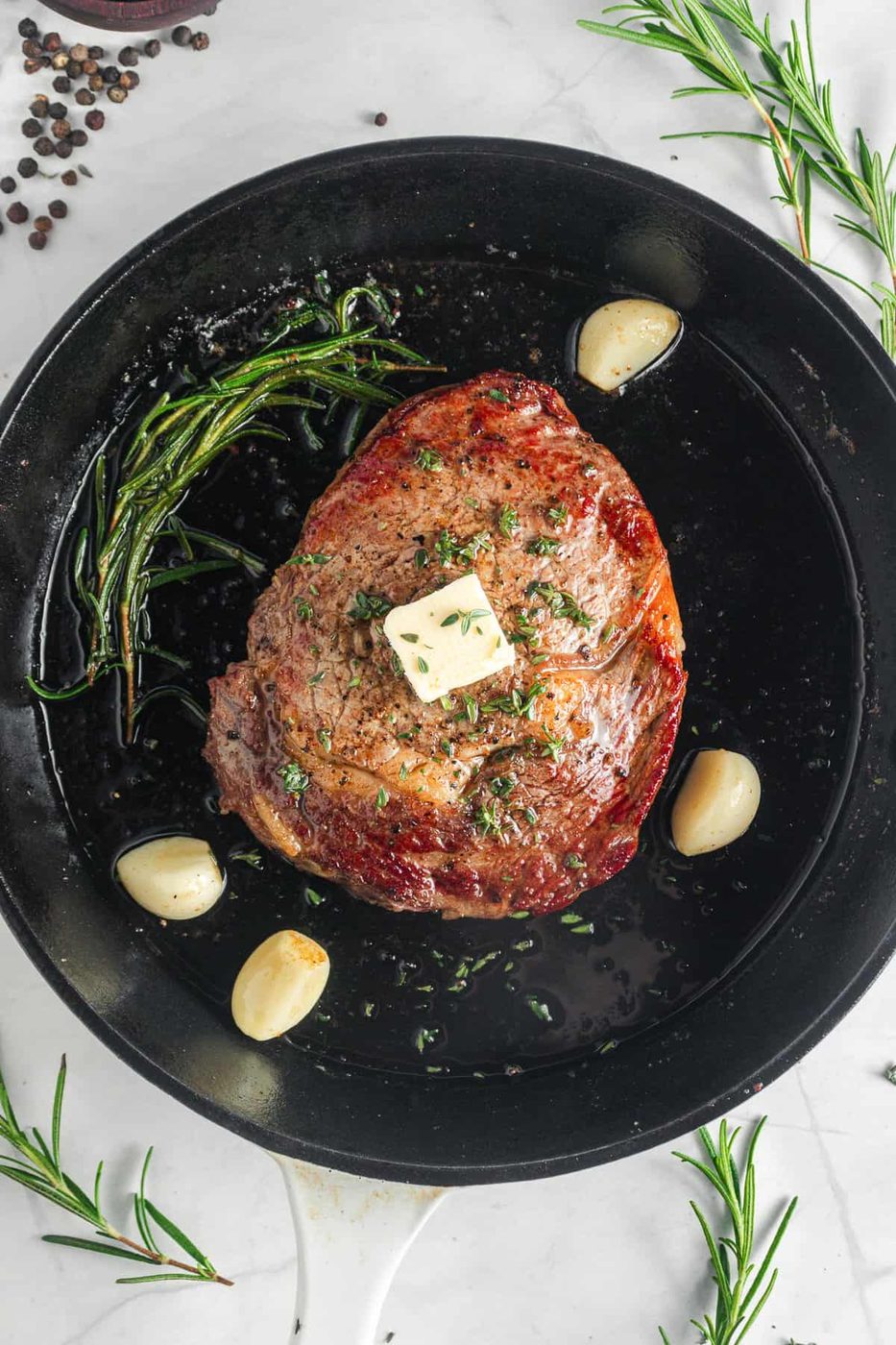 The grilled meat topped with a pat of butter and sprinkled with herbs sits in a skillet. Surrounding the meat are cloves of garlic and sprigs of rosemary.
