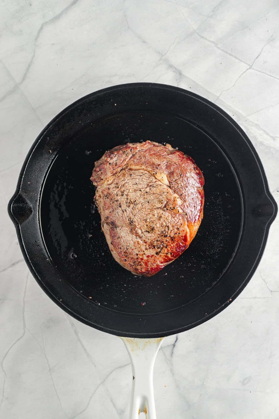 A piece of seared meat cooking in a black cast iron skillet on a gray marble surface. The meat has a browned exterior, and the pan shows traces of oil.