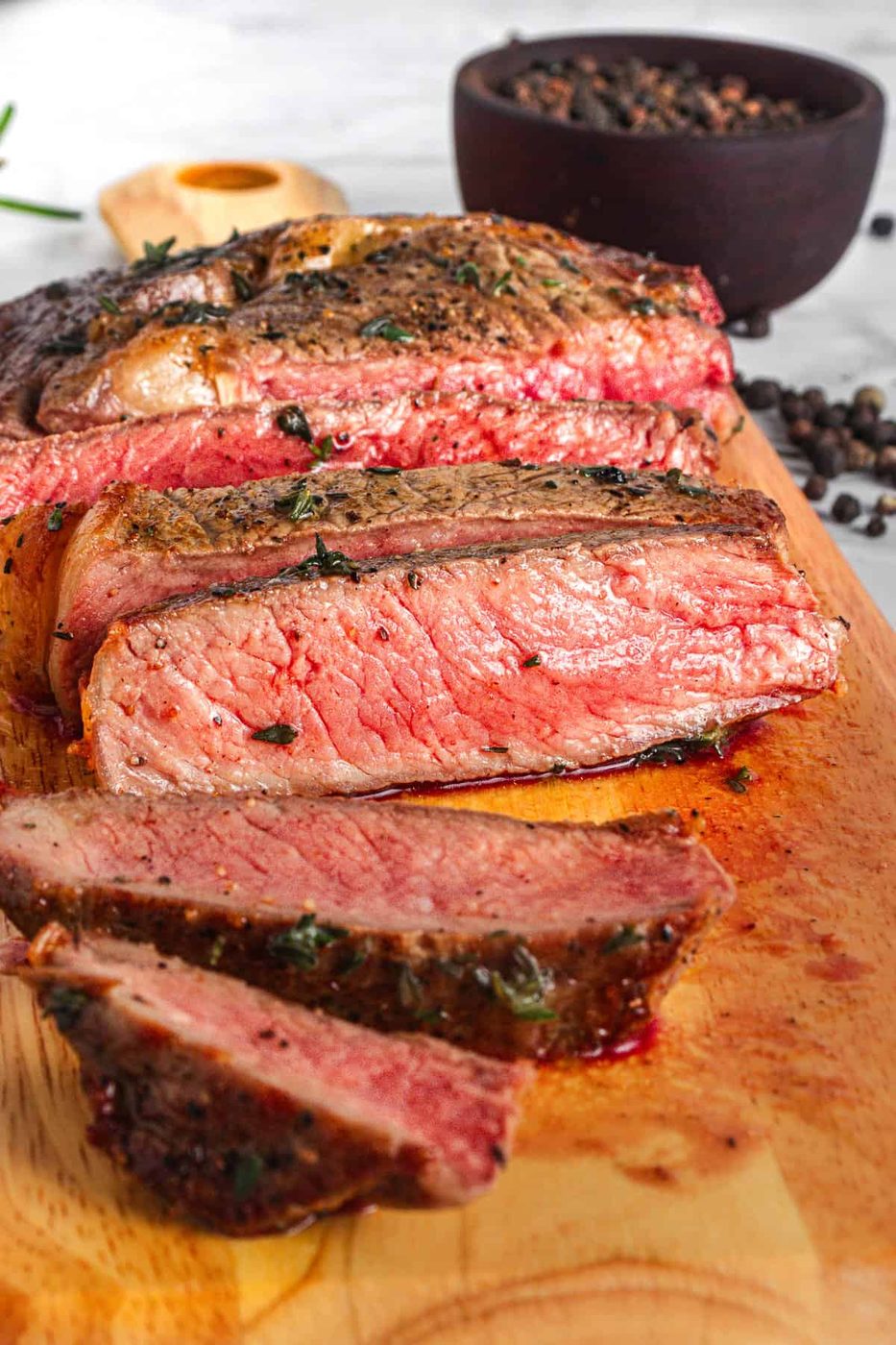 Slices of medium-rare steak on a wooden cutting board, garnished with herbs. In the background, a black bowl filled with peppercorns and a wooden spoon are visible. The steak is juicy with a seared crust and pink interior.