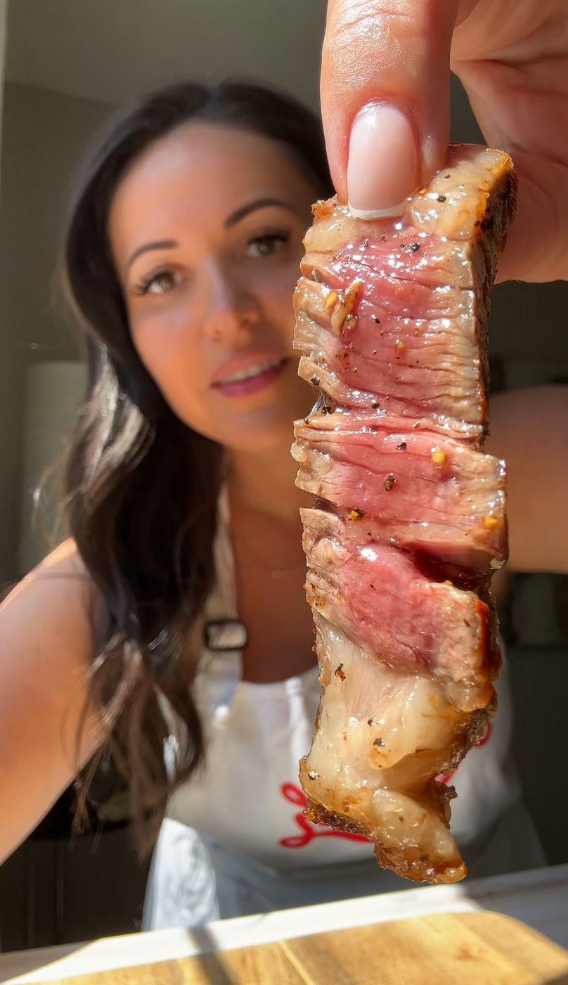A woman holds up a juicy, grilled steak slice, showing the medium-rare interior. She is smiling and wearing a white apron. The background is softly blurred, highlighting the focus on the steak.