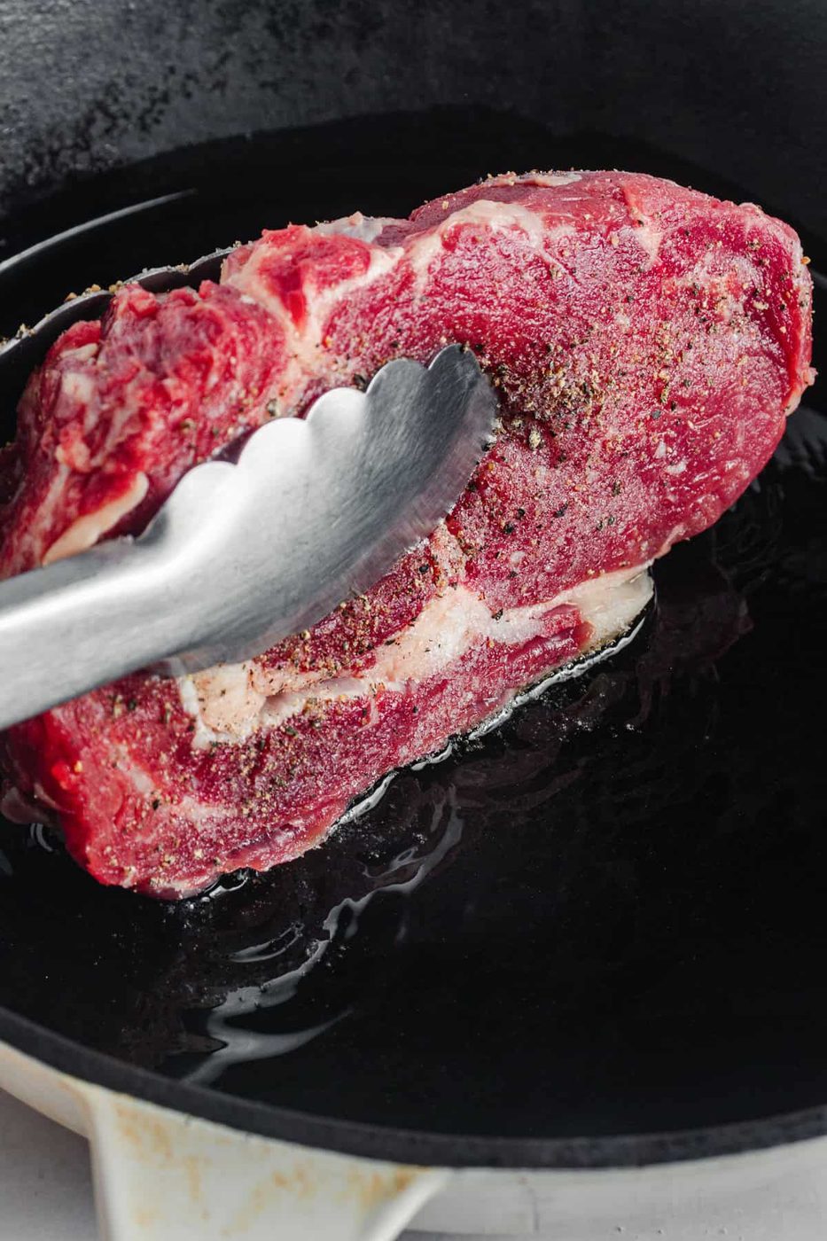 A raw, marbled steak seasoned with pepper is being placed into a hot cast iron skillet using metal tongs. The steak appears to be in the initial stages of cooking.