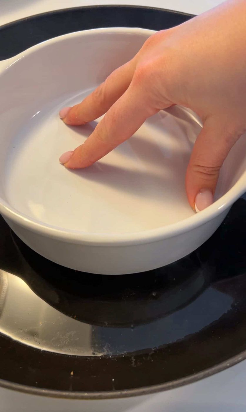 A hand with light pink nails is gently touching the inside of a white ceramic dish, which is placed on a black stovetop.
