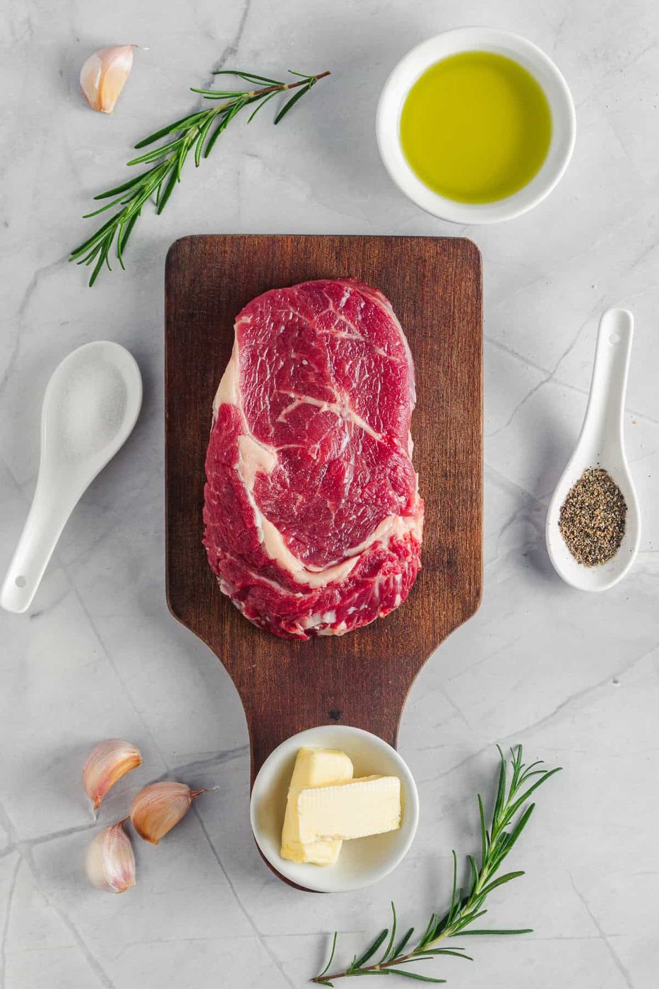 A raw steak on a wooden cutting board surrounded by small bowls of olive oil, butter, garlic cloves, rosemary sprigs, and spoons of salt and pepper, all placed on a light marble surface.