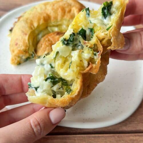 A hand holds a flaky pastry filled with creamy cheese and spinach. Another portion of the same pastry sits on a white plate, all on a wooden table background.