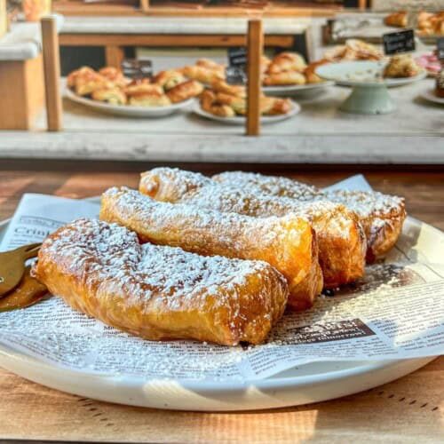A plate of golden, flaky pastries dusted with powdered sugar sits on a newspaper-lined dish, with more baked goods and pastries visible in the blurred background of a bakery display.