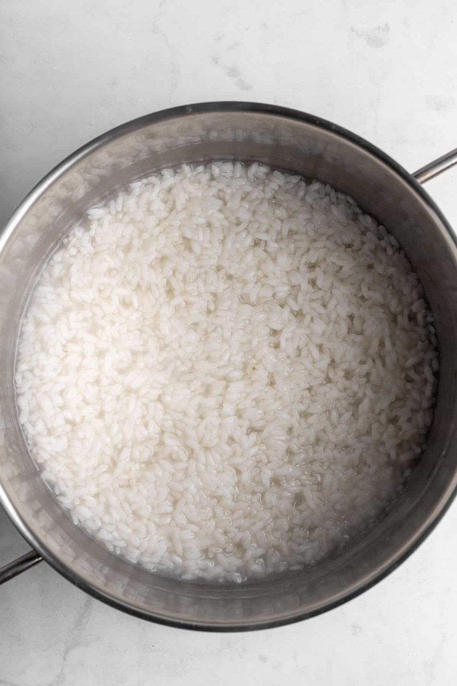 A pot filled with cooked white rice sits on a marble surface. The rice appears soft and slightly sticky, evenly spread across the flat surface of the pot. The metal handles of the pot are visible on the sides.