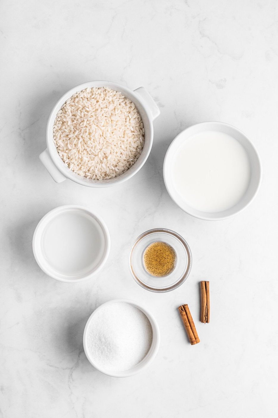 Top view of ingredients for rice pudding on a marble surface. The items include a bowl of white rice, a bowl of milk, a bowl of water, a small bowl of sugar, a small bowl of ground cinnamon, and two cinnamon sticks.