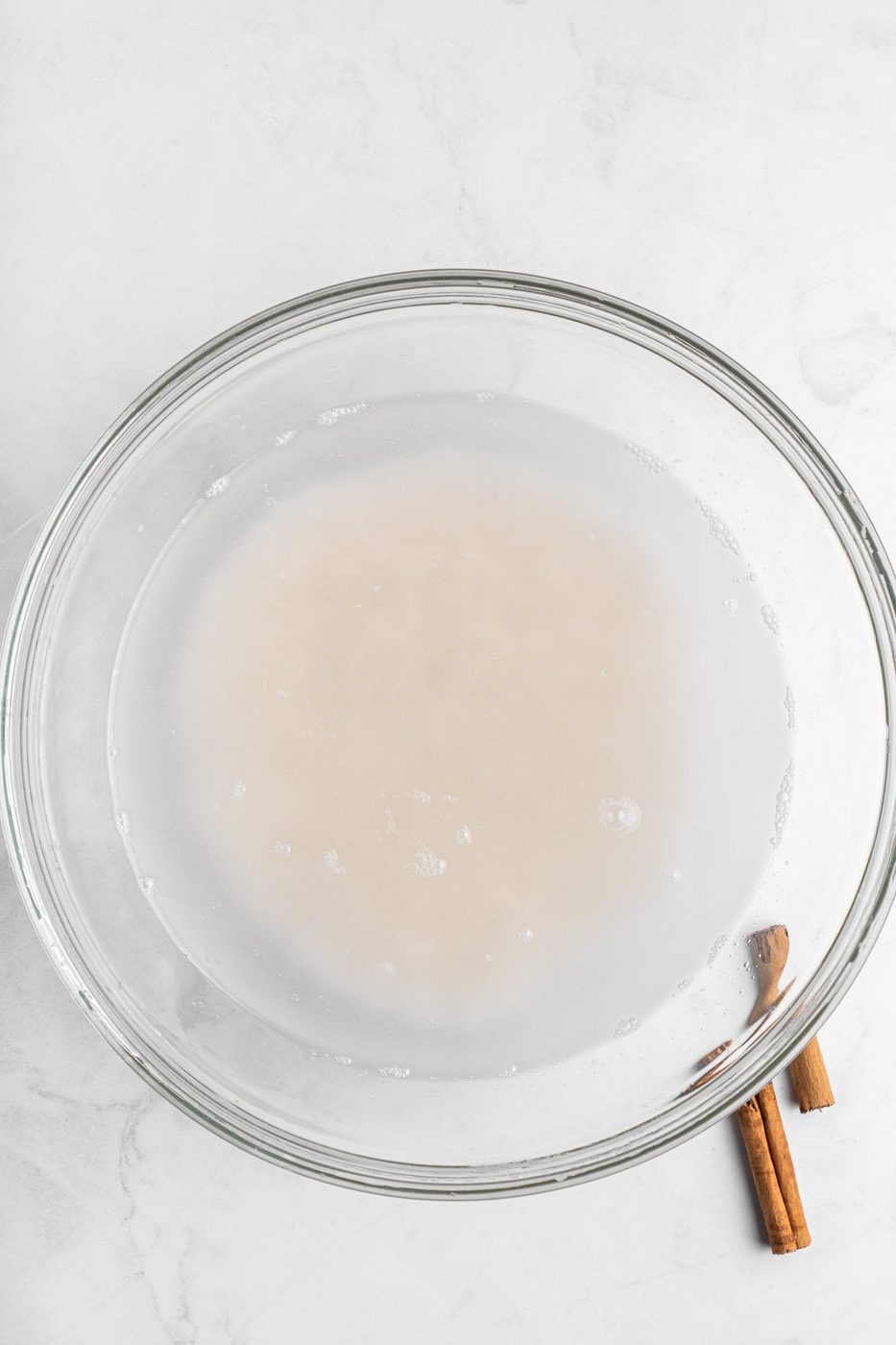 A clear glass bowl contains a light milky liquid on a white marble surface. Two cinnamon sticks are placed beside the bowl on the right.